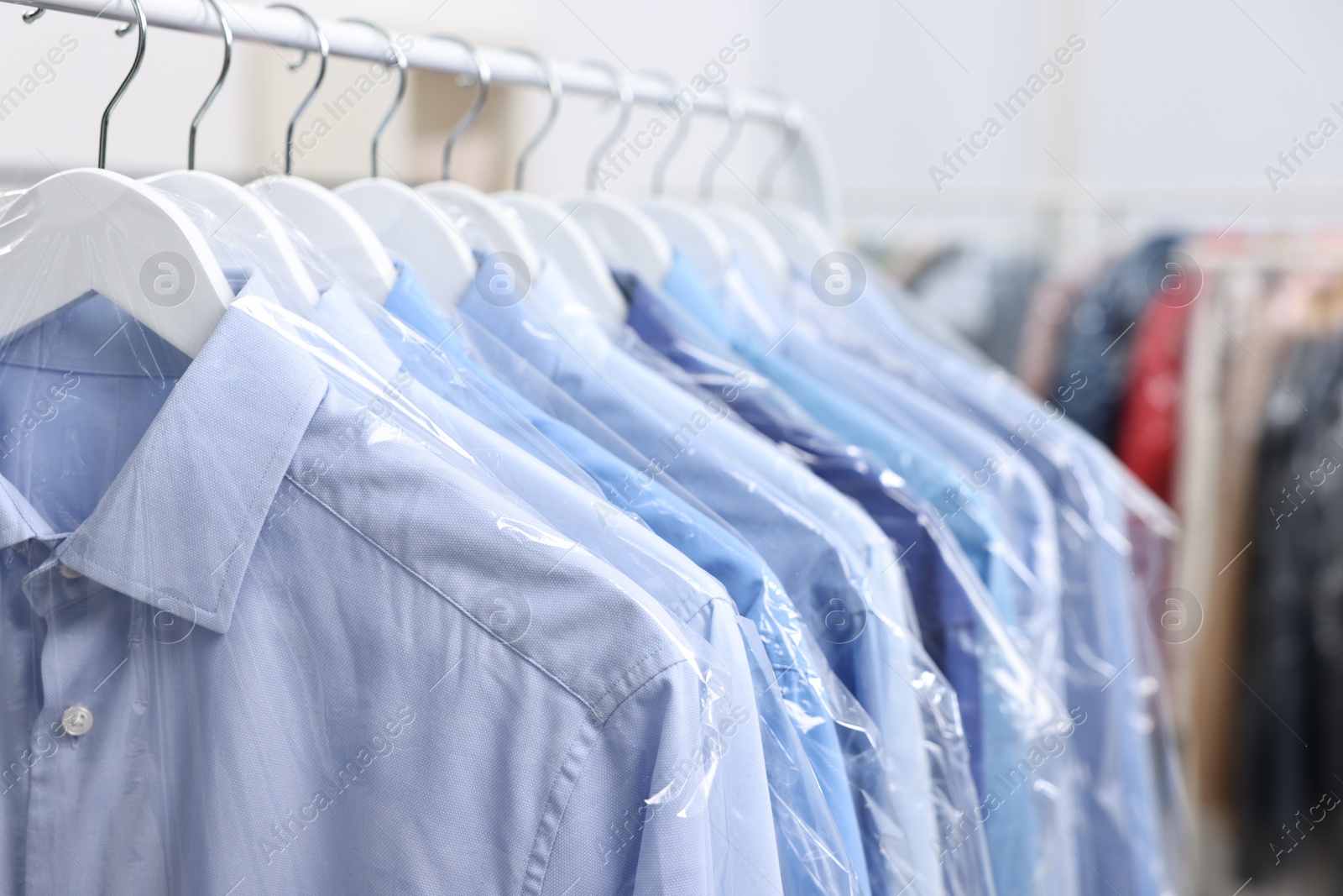 Photo of Dry-cleaning service. Many different clothes in plastic bags hanging on rack indoors, closeup