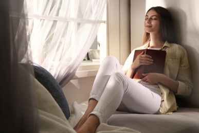 Photo of Beautiful young woman with book relaxing near window at home