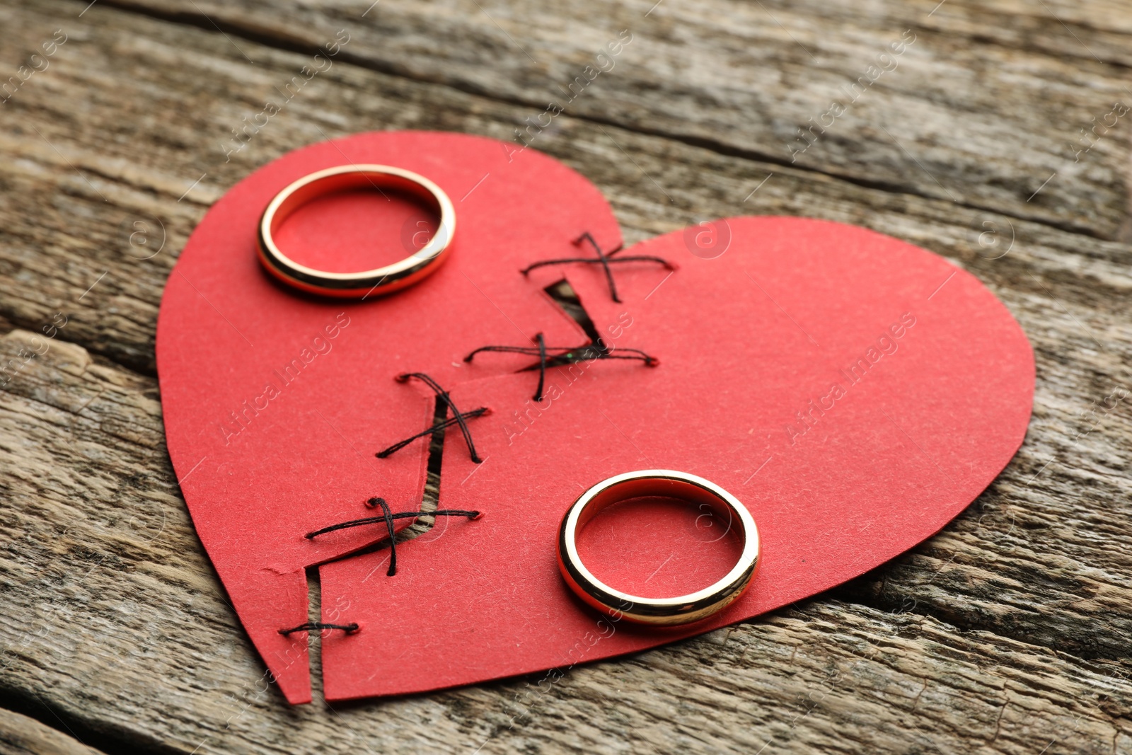 Photo of Broken heart. Torn red paper heart sewed with thread and wedding rings on wooden table, closeup