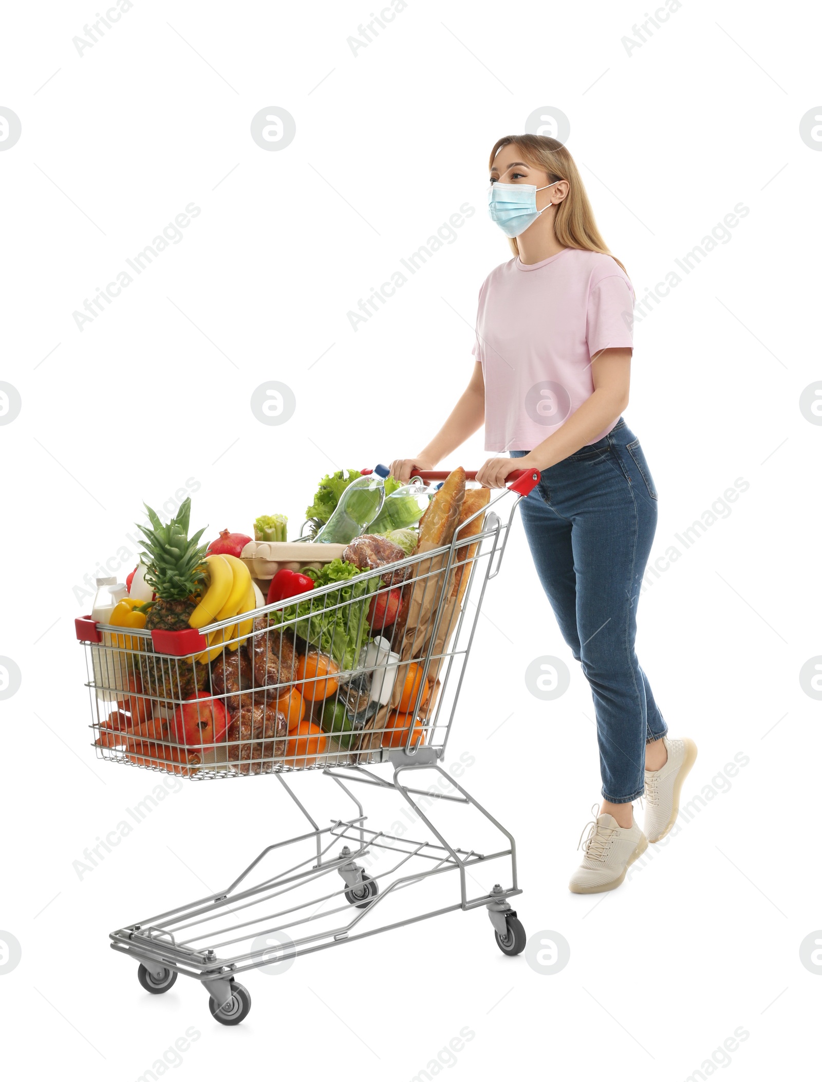 Photo of Young woman in medical mask with shopping cart full of groceries on white background