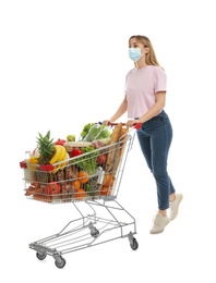 Young woman in medical mask with shopping cart full of groceries on white background