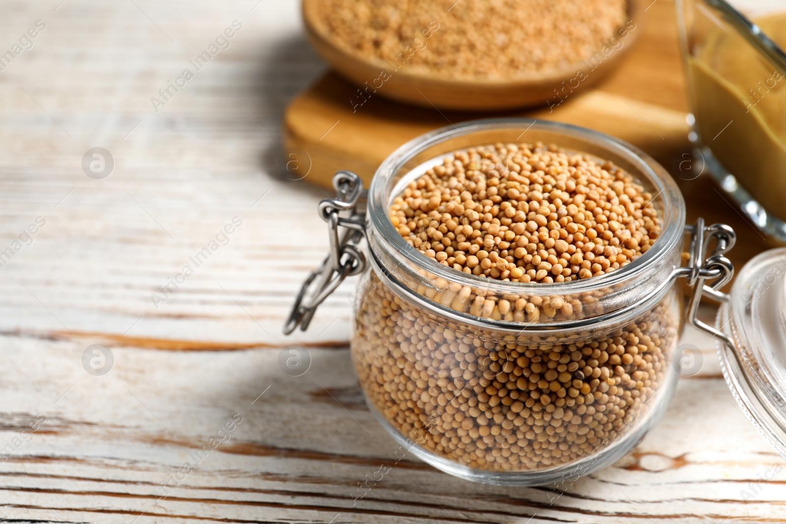 Photo of Mustard seeds in glass jar on white wooden table, closeup. Space for text