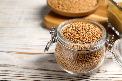 Photo of Mustard seeds in glass jar on white wooden table, closeup. Space for text