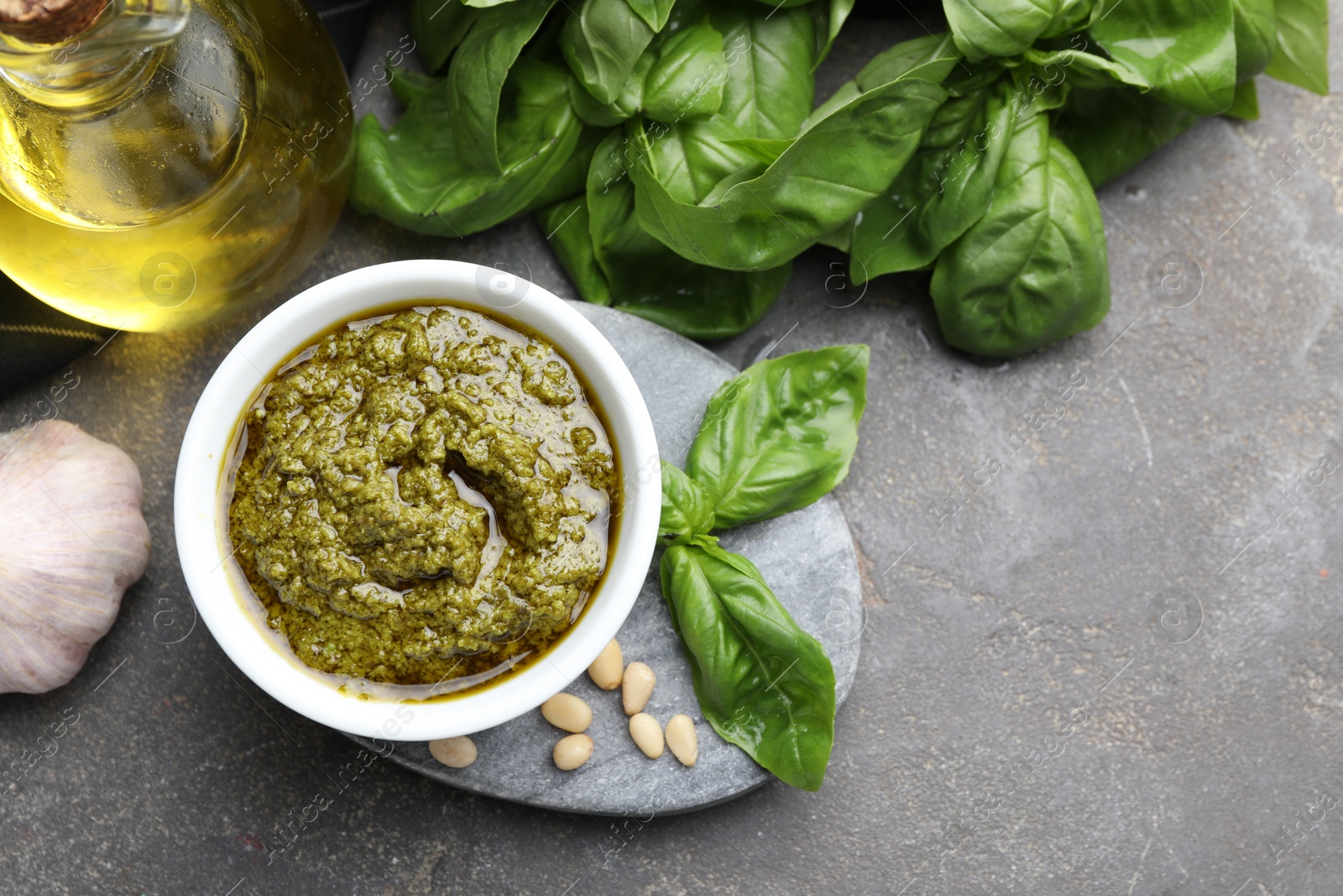 Photo of Tasty pesto sauce in bowl, basil, pine nuts, garlic and oil on grey table, top view. Space for text