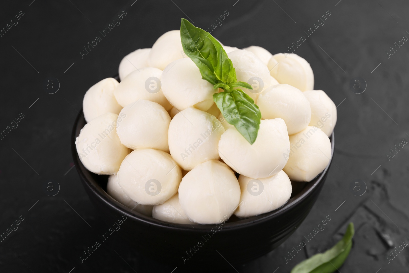 Photo of Tasty mozzarella balls and basil leaves in bowl on black table, closeup