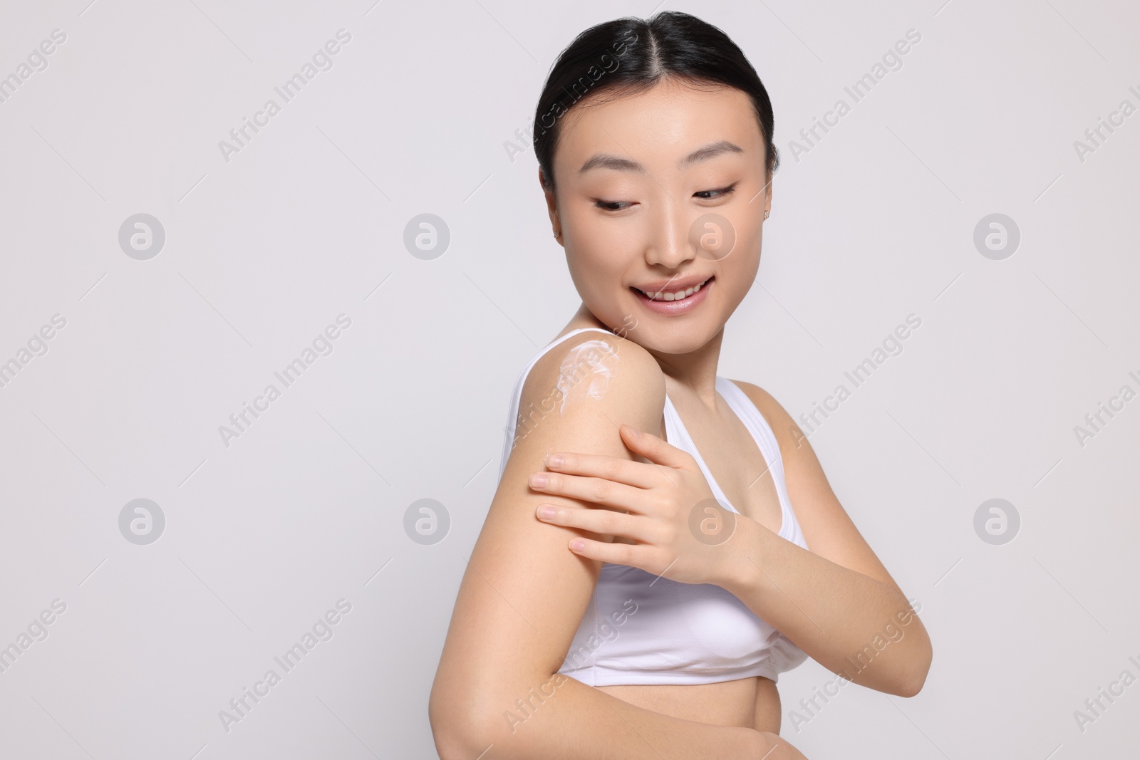 Photo of Beautiful young Asian woman applying body cream onto shoulder on light grey background, space for text