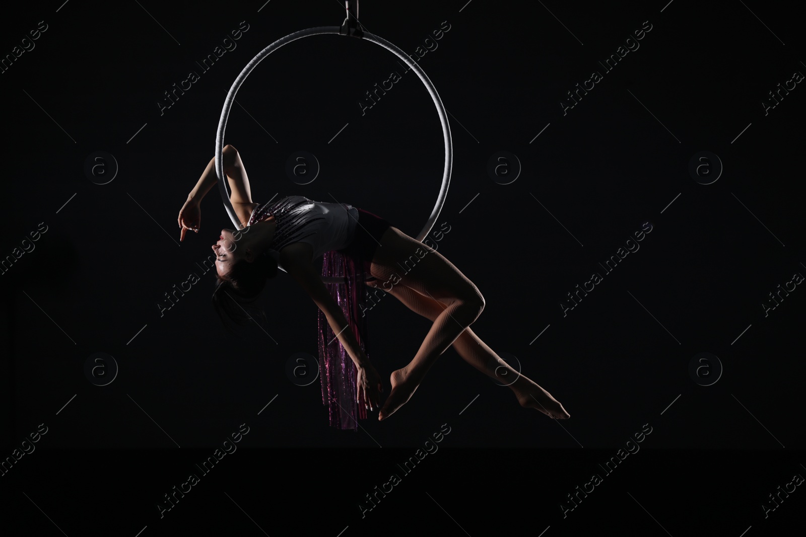 Photo of Young woman performing acrobatic element on aerial ring against dark background