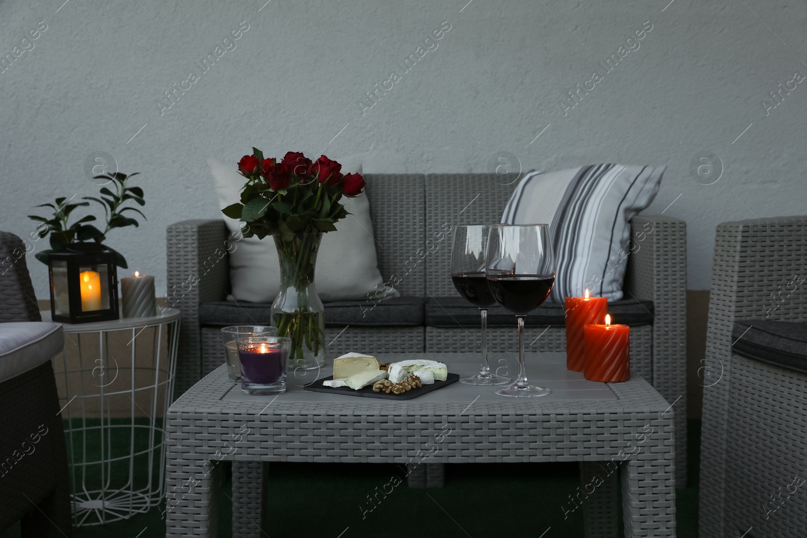 Photo of Glasses of wine, vase with roses, burning candles and snacks on rattan table at balcony in evening