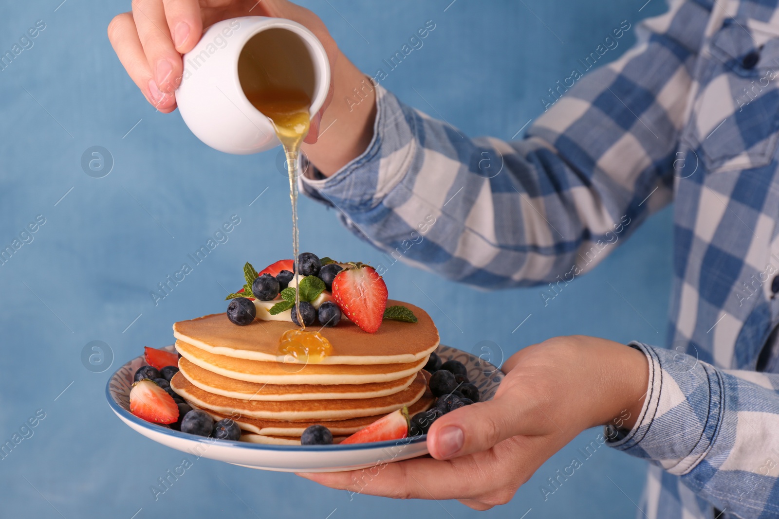 Photo of Woman pouring honey onto delicious pancakes with fresh berries and butter against light blue background, closeup