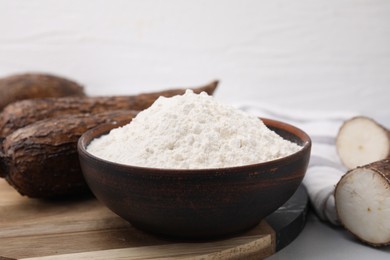Photo of Bowl with cassava flour and roots on white table, closeup