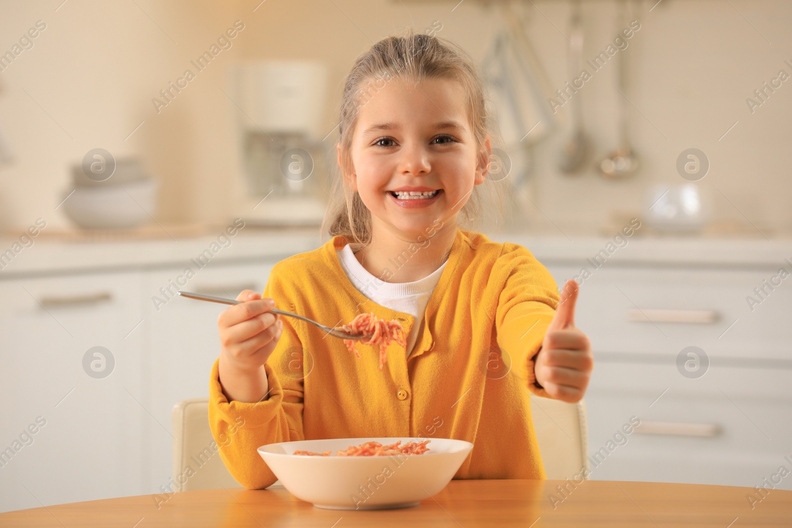 Photo of Cute little girl eating tasty pasta at table in kitchen