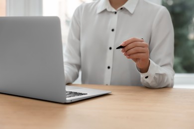 Woman with pen working on laptop at wooden table, closeup. Electronic document management