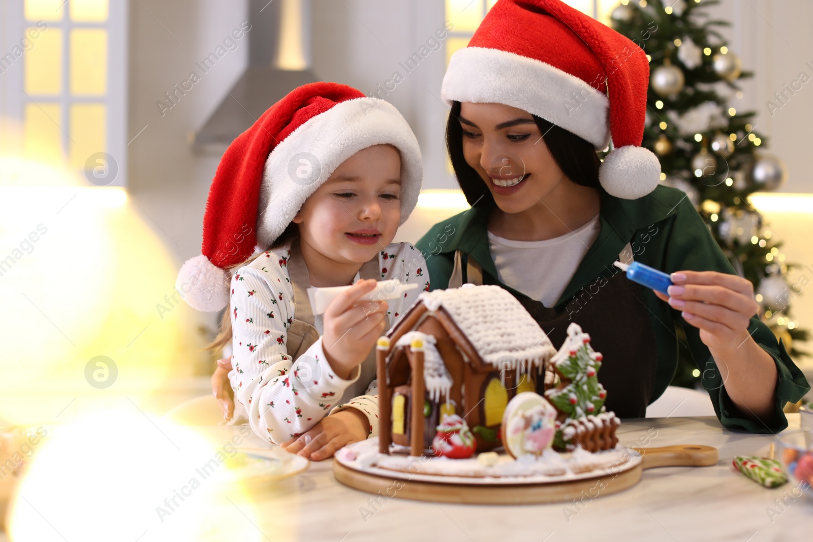 Photo of Mother and daughter decorating gingerbread house at table indoors