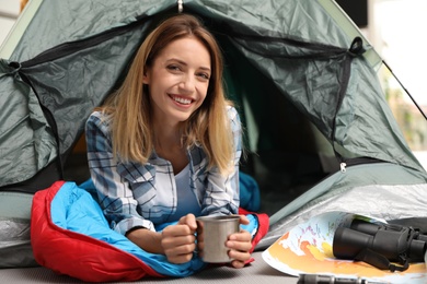 Young woman in sleeping bag with mug looking outside of tent