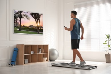 Image of Sporty man with remote control training on walking treadmill and watching TV at home