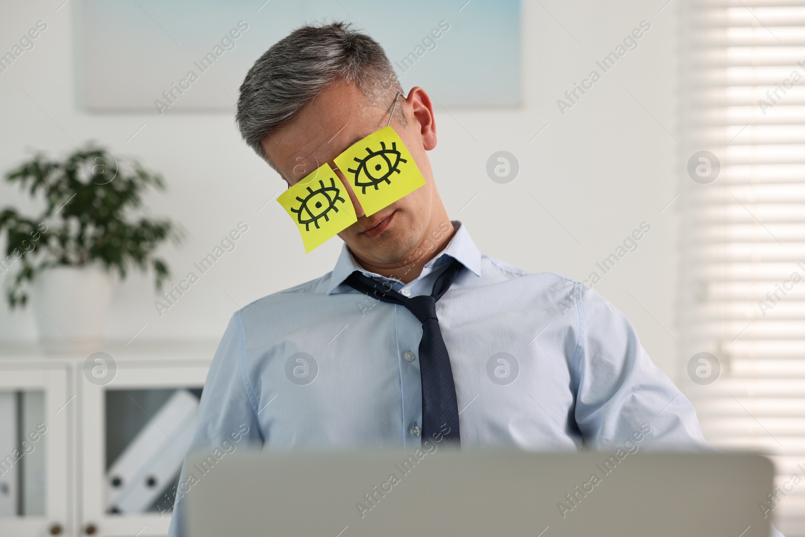 Photo of Man with fake eyes painted on sticky notes snoozing at workplace in office