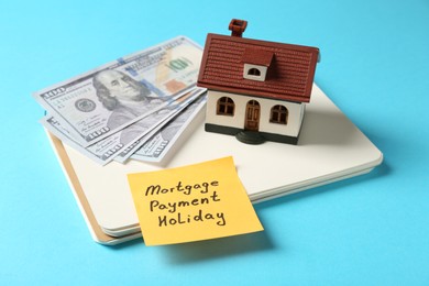 Photo of Paper note with words Mortgage Payment Holiday, notebook, house model and money on light blue background
