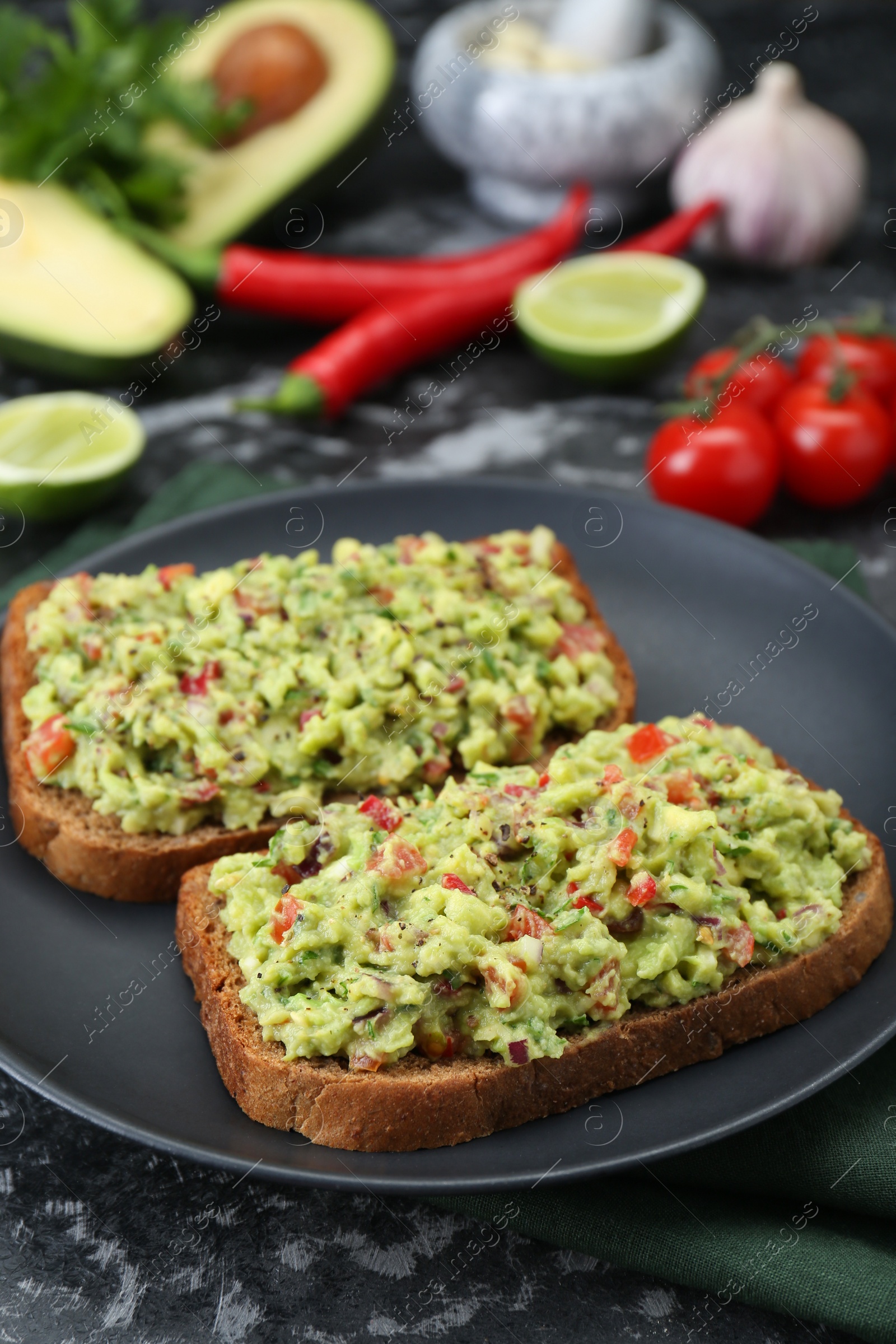 Photo of Slices of bread with tasty guacamole and ingredients on black textured table, closeup