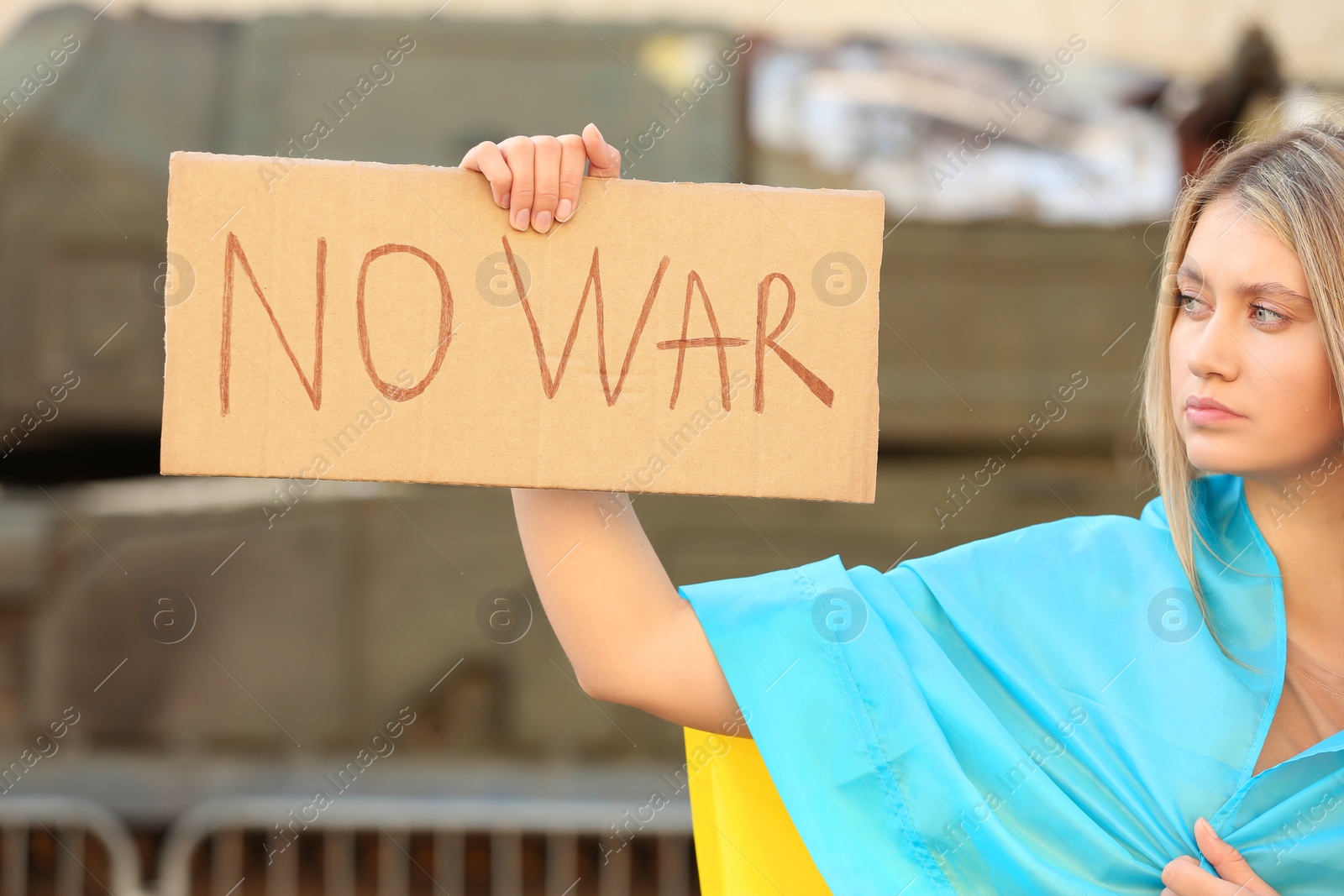 Photo of Sad woman wrapped in Ukrainian flag holding poster with words No War outdoors