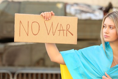 Photo of Sad woman wrapped in Ukrainian flag holding poster with words No War outdoors