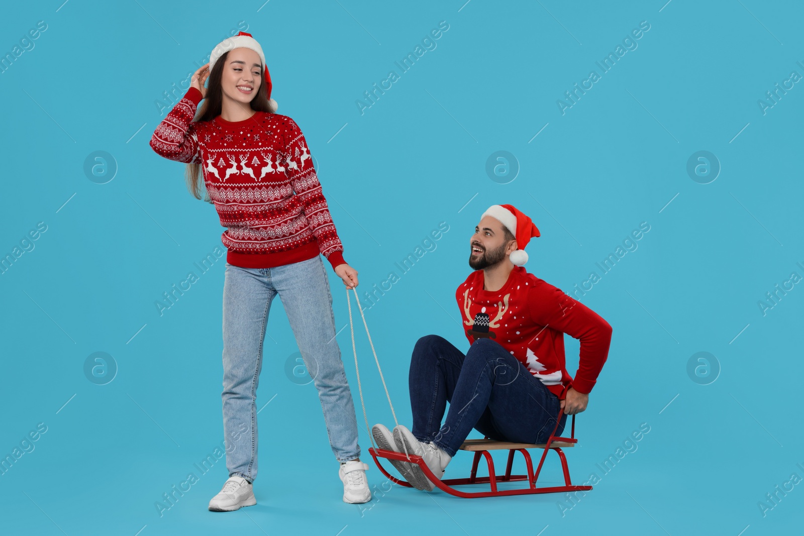 Photo of Young couple in Christmas sweaters. Woman pulling her man in sled on light blue background