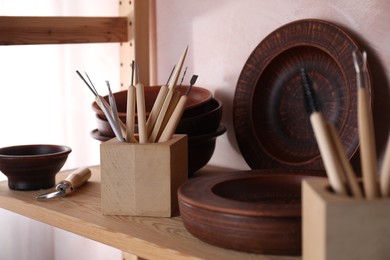 Set of different crafting tools and clay dishes on wooden rack in workshop