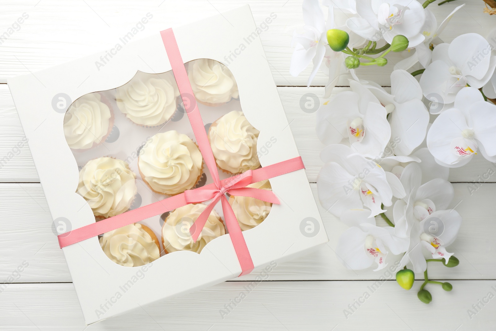 Photo of Tasty cupcakes with vanilla cream in box and orchid flowers on white wooden table, top view