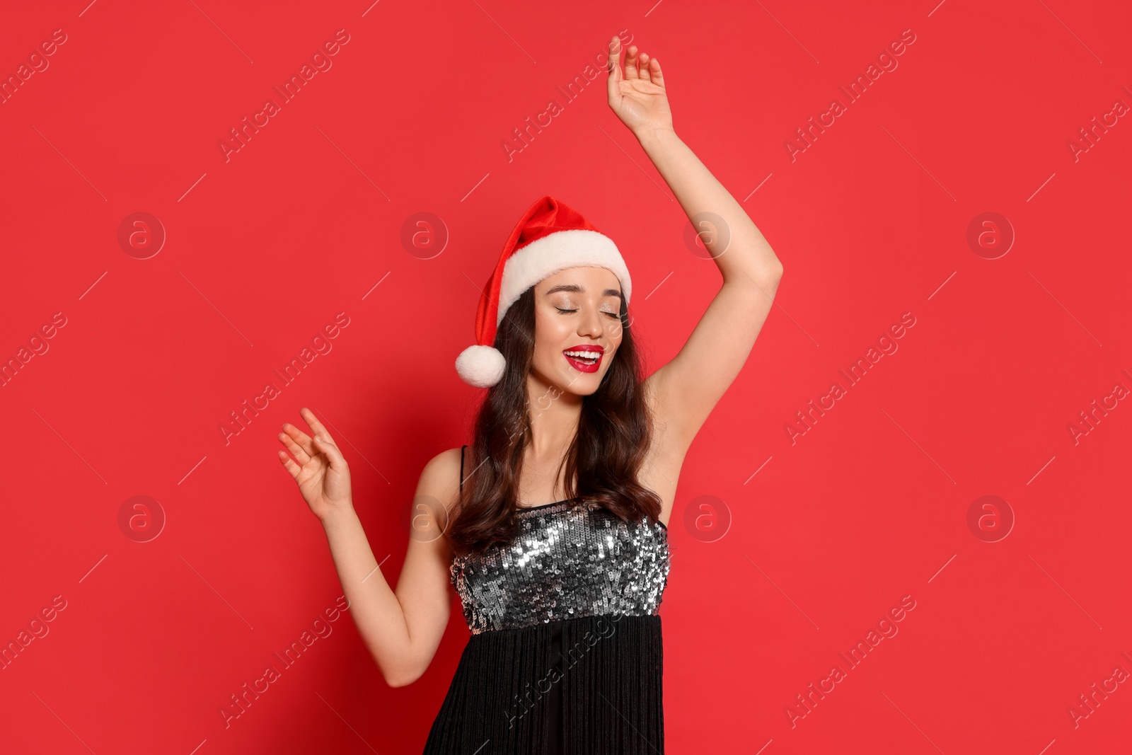 Photo of Christmas celebration. Beautiful young woman in stylish dress and Santa hat on red background