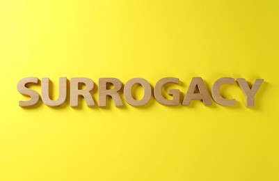 Photo of Word Surrogacy made of wooden letters on yellow background, flat lay