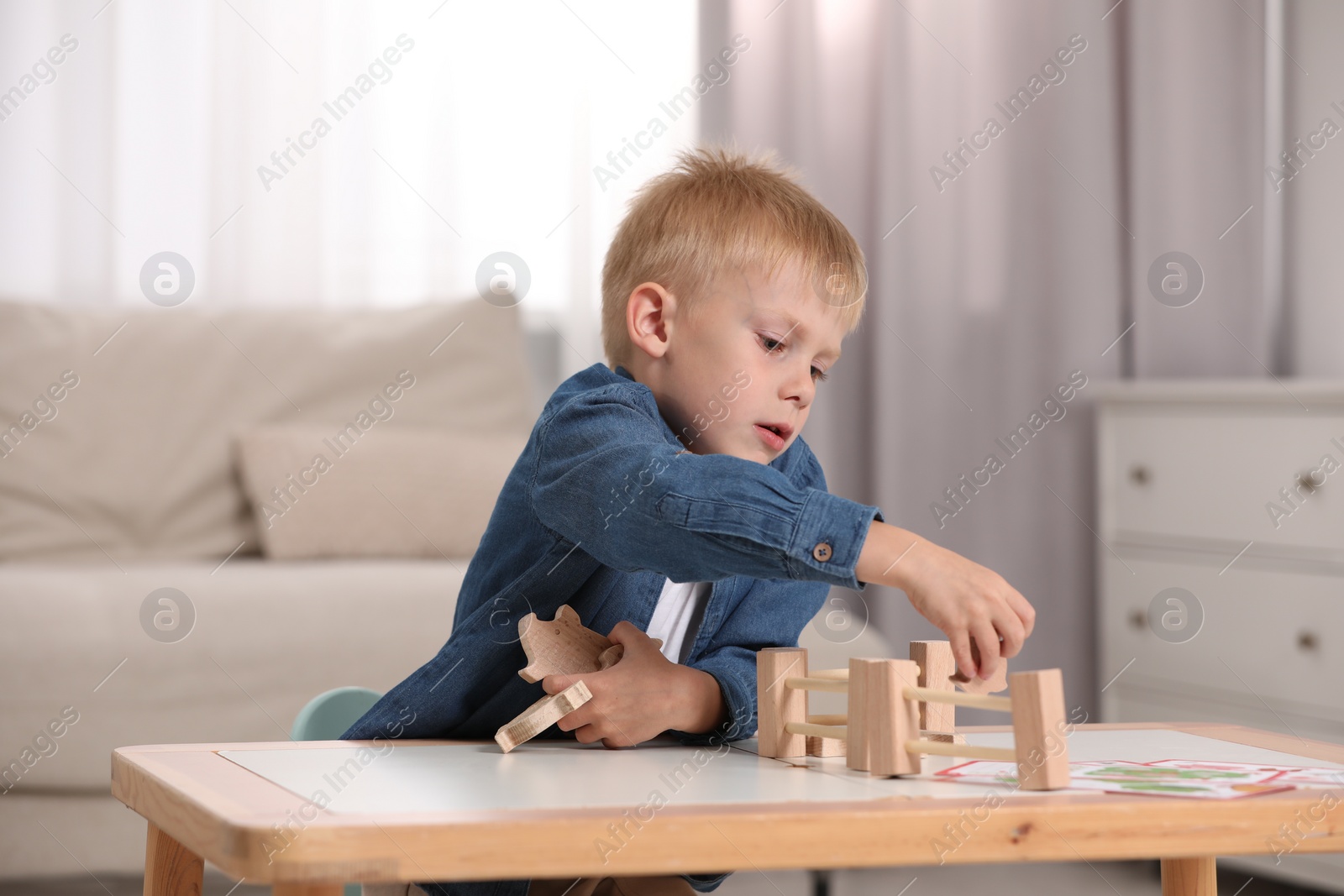 Photo of Cute little boy playing with set of wooden animals and fence at table indoors. Child's toy