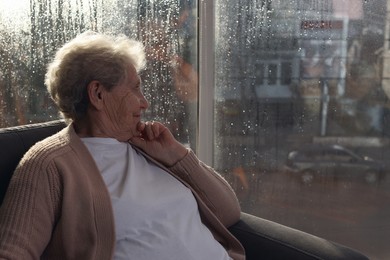 Photo of Elderly woman looking out of window on rainy day, space for text. Loneliness concept