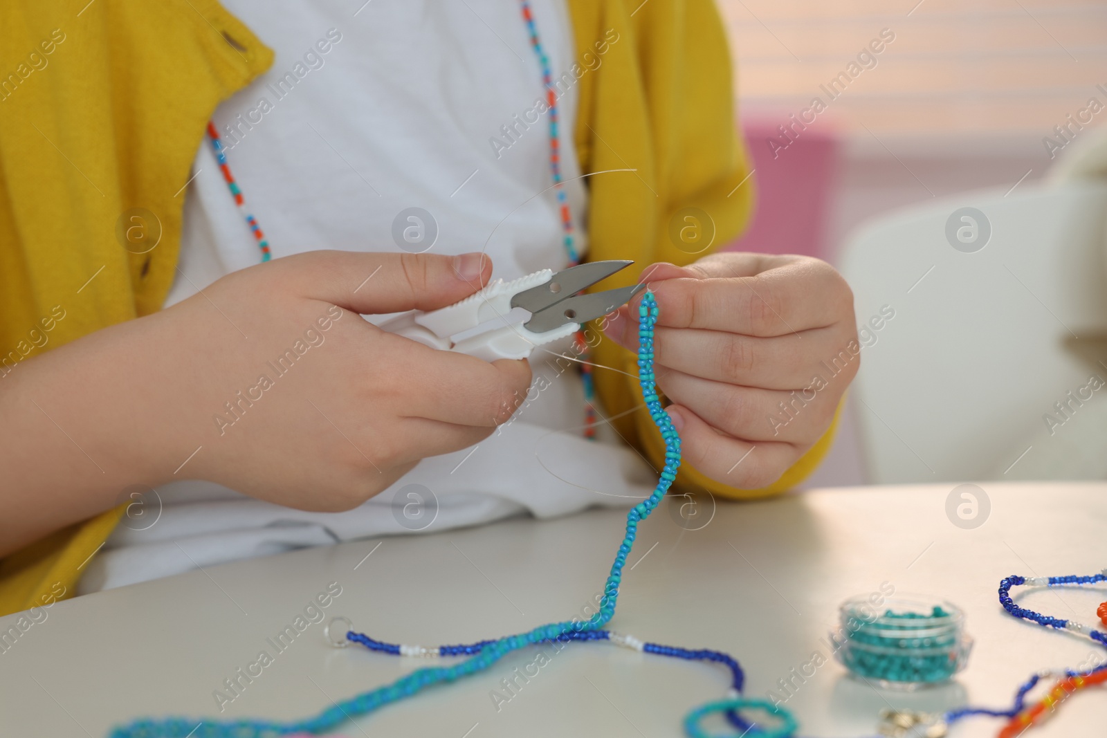 Photo of Little girl making beaded jewelry at table in room, closeup