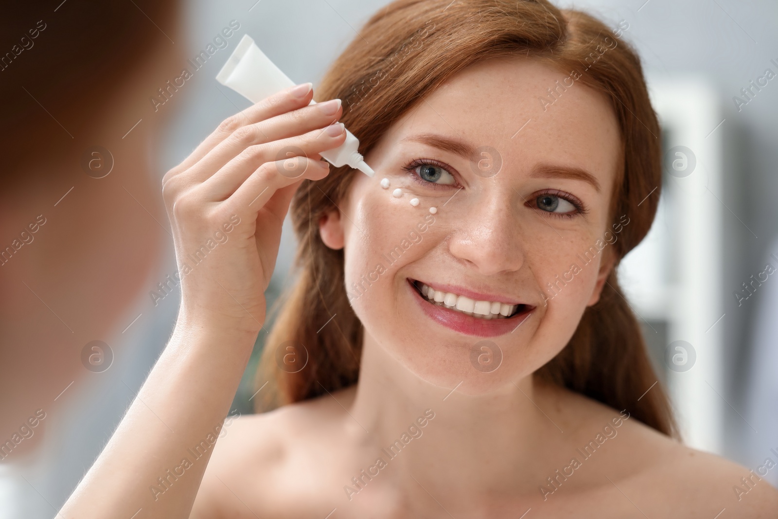 Photo of Smiling woman with freckles applying cream onto her face near mirror in bathroom