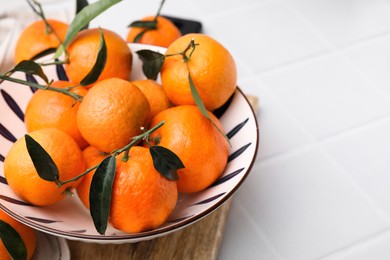 Photo of Fresh ripe tangerines with green leaves in bowl on white tiled table, closeup. Space for text