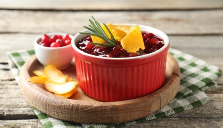 Photo of Cranberry sauce in bowl, fresh berries, rosemary and orange peels on wooden table, closeup