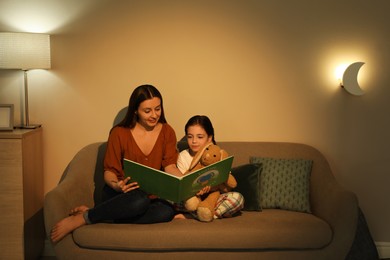 Photo of Little girl with mother reading book in living room lit by night lamp