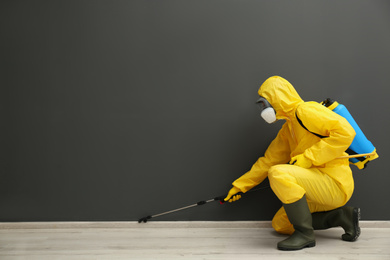 Pest control worker in protective suit spraying pesticide near black wall indoors. Space for text