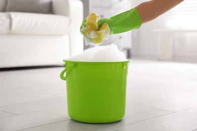 Photo of Woman holding sponge with foam over bucket indoors, closeup. Cleaning supplies