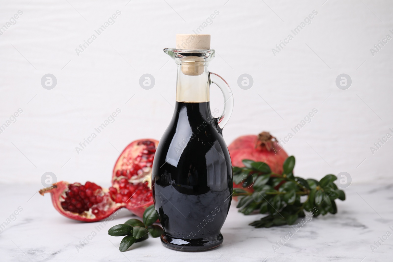 Photo of Tasty pomegranate sauce in bottle, fruits and branches on white marble table, closeup