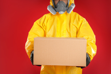 Photo of Man wearing chemical protective suit with cardboard box on red background, closeup. Prevention of virus spread