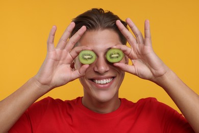Photo of Smiling man covering eyes with halves of kiwi on orange background, closeup