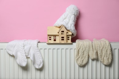 Photo of Modern radiator with knitted hat, socks, mittens and wooden house near pink wall indoors. Winter heating efficiency