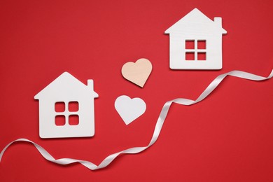 Photo of White ribbon and decorative hearts between two house models symbolizing connection in long-distance relationship on red background, flat lay