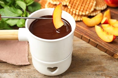 Photo of Dipping slice of peach into fondue pot with chocolate on wooden table, closeup