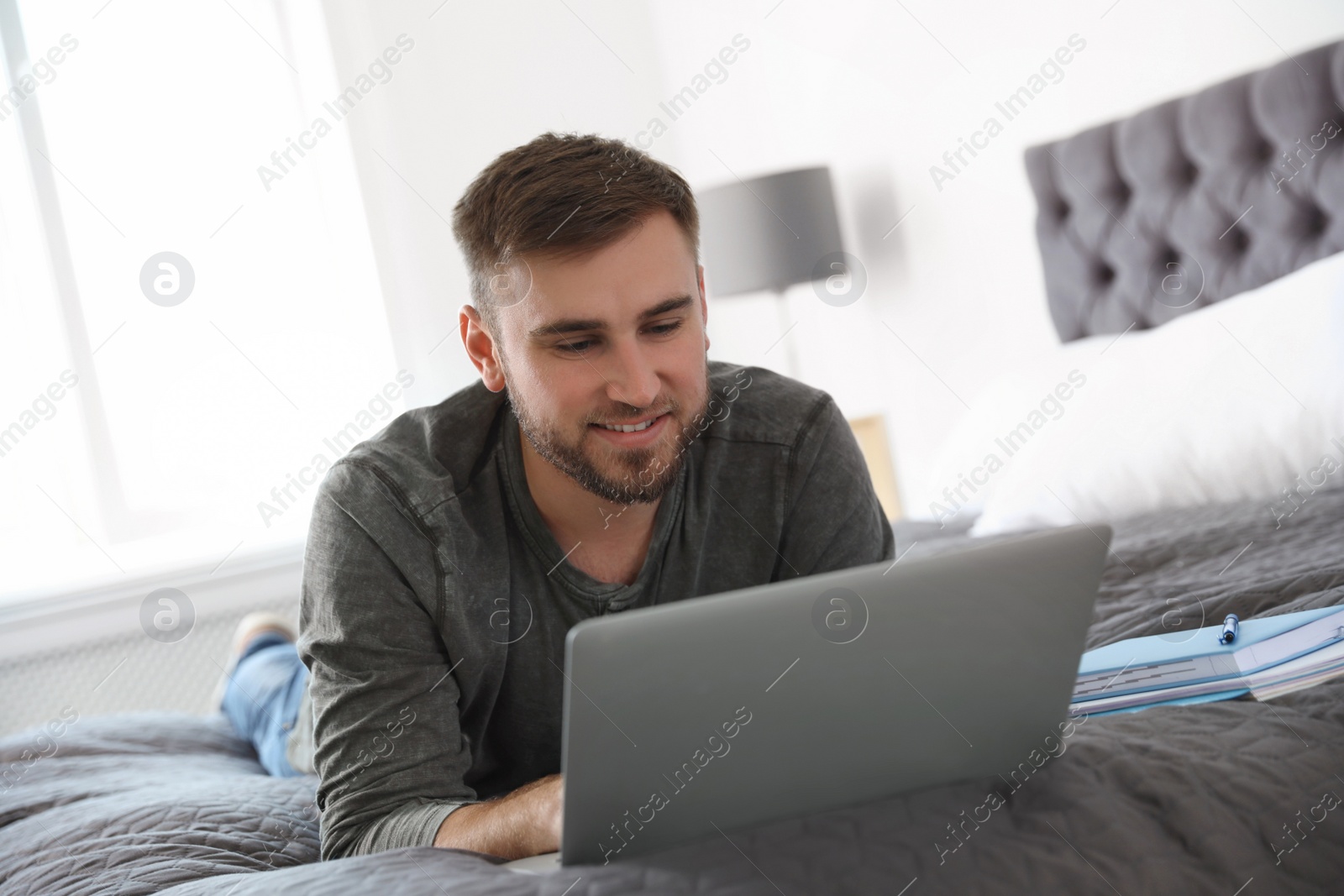 Photo of Young man using laptop while lying on bed at home