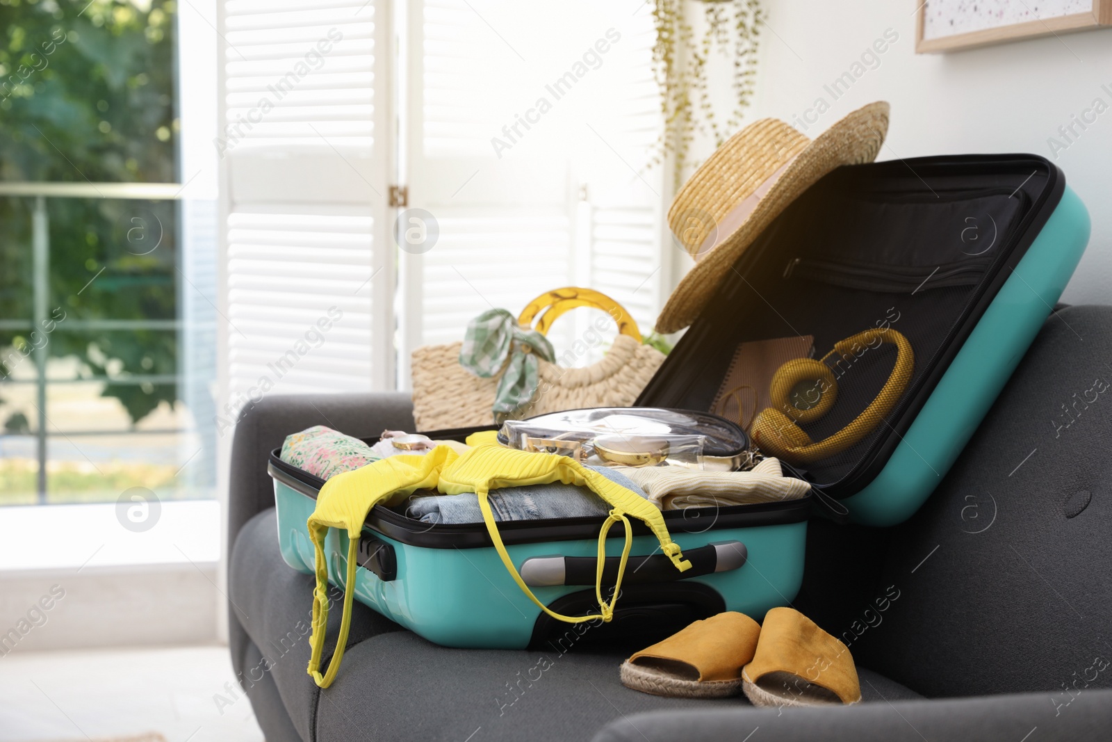 Photo of Open suitcase full of clothes, shoes and summer accessories on sofa in room