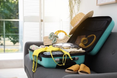 Open suitcase full of clothes, shoes and summer accessories on sofa in room