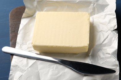 Photo of Block of tasty butter in open foil packaging and knife on blue wooden table, closeup