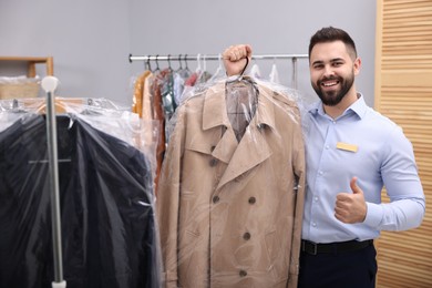 Photo of Dry-cleaning service. Happy worker holding hanger with coat in plastic bag and showing thumb up indoors
