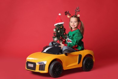 Photo of Cute little girl with Christmas tree driving children's electric toy car on red background